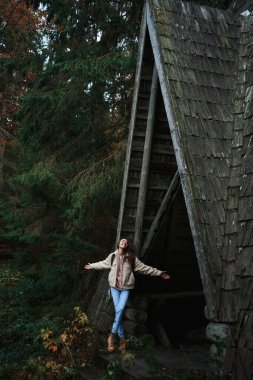 Overjoyed hipster girl posing with open arms by vintage style wooden hut during travel in Carpathian Ukraine, following to insta place