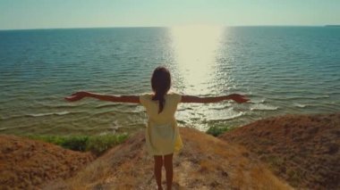 back view happy woman in yellow dress walks on edge of clay hill over sea with morning sunlight. travel in Ukraine, Stanislav cliffs, Kherson region