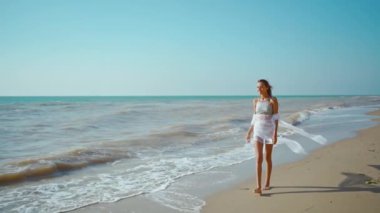 Ağır çekim yaz ve seyahat konsepti. Kumsalda pareo ile bikinili mutlu bir genç kadın. Neşeli, ince vücutlu kız yürüyor rüzgar ve dalgaların tadını çıkarıyor..