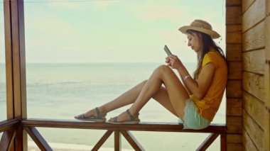 Plajda şapkalı bir kadın, akıllı telefon kullanıyor, internette sohbet ediyor, internette geziniyor. Teknoloji ve seyahat, tatilde online iletişim..