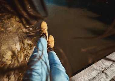 Nehir suyunun üzerinde kadın bacakları, kahverengi saçlar kamerayı çerçeveler. Kız tahta köprüde oturuyor.