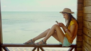 Sahilde şapkalı bir kadın dizüstü bilgisayar kullanıyor, akıllı telefon kullanıyor, internette sohbet ediyor, internette geziniyor. Teknoloji ve seyahat, tatilde online iletişim..