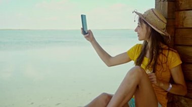 Sahilde şapkalı bir kadın dizüstü bilgisayar kullanıyor, akıllı telefon kullanıyor, internette sohbet ediyor, internette geziniyor. Teknoloji ve seyahat, tatilde online iletişim..