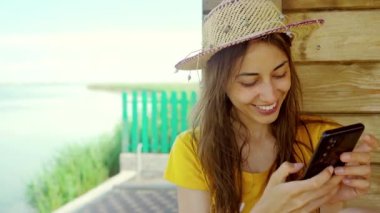 Sahilde şapkalı bir kadın dizüstü bilgisayar kullanıyor, akıllı telefon kullanıyor, internette sohbet ediyor, internette geziniyor. Teknoloji ve seyahat, tatilde online iletişim..