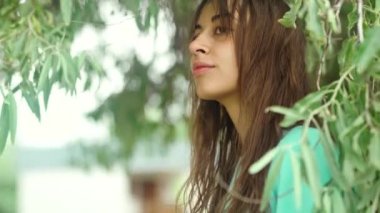 Kameraya gülümseyen ve yeşil zeytin ağacının önünde mutlu görünen çekici esmer kadının samimi portresi. Turkuazlı kız yapraklarla oynuyor. Doğal görünüm kavramı