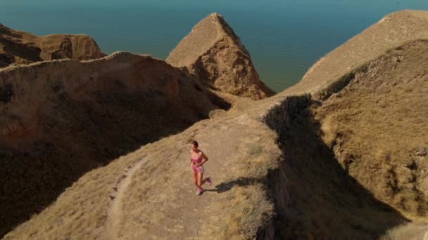 フィットネスの女性ジョガーの空中スローモーション映像海の海岸の美しい粘土の峡谷に沿って早朝の屋外で実行しています 夏の日の出に運動をする 健康的なライフスタイルとスポーツ — ストック動画