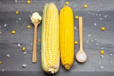 Tahta bir kaşıkta tuz ve tereyağı ile çevrili gri bir masada iki taze ve pişmiş mısır koçanı yatıyor..