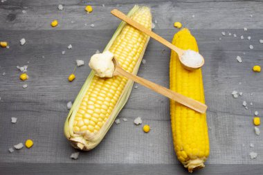 Tahta bir kaşıkta tuz ve tereyağı ile çevrili gri bir masada iki taze ve pişmiş mısır koçanı yatıyor..