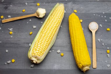 Tahta bir kaşıkta tuz ve tereyağı ile çevrili gri bir masada iki taze ve pişmiş mısır koçanı yatıyor..