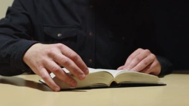 Koyu renk gömlekli, kimliği belirsiz bir adamın elleri kalın bir kitabın çarşaflarını çeviriyor.