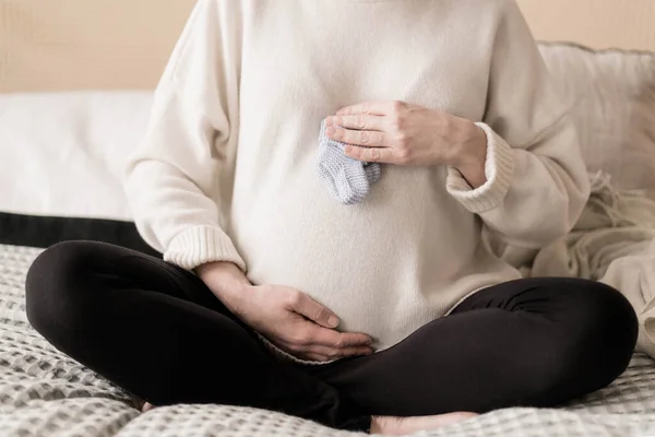 Una mujer embarazada está sentada en la cama en un suéter caliente y sosteniendo su vientre. — Foto de Stock