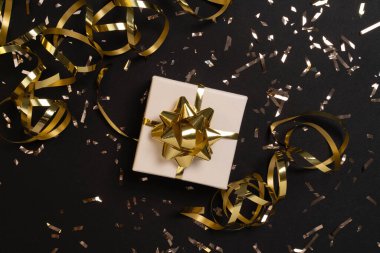Altın yaylı küçük beyaz hediye kutusu ve siyah arka planda altın konfeti.