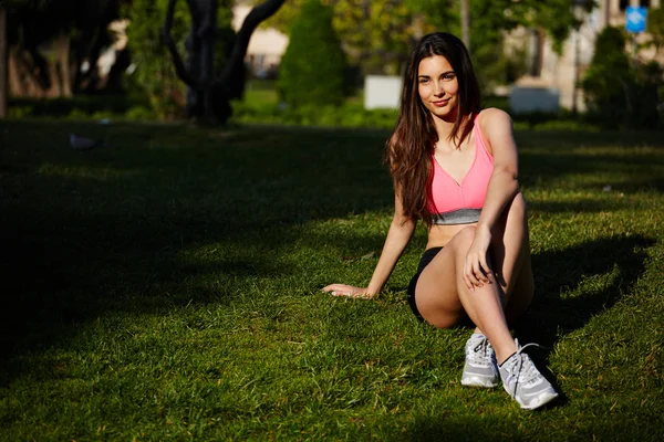 Athletic girl seated on the grass and resting after the jogging in the beautiful park — Stock Photo, Image