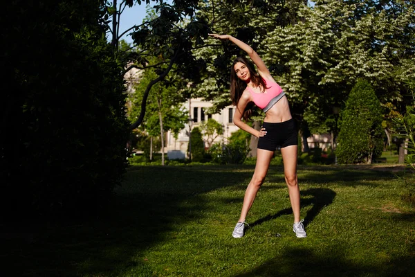 运动女孩做伸展运动，站在美丽的公园里的草地上 — 图库照片