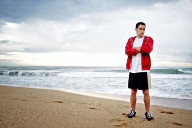 genç sporcu adam beyaz tişört ve spor kumun üzerinde duran ve güzel deniz keyfi kırmızı bir yağmurluk