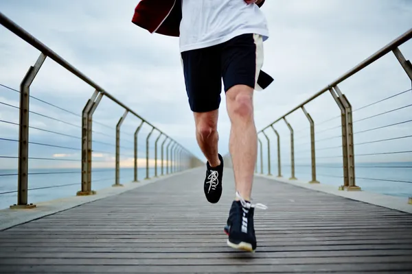 Young athlete man in white t-shirt and sports a red windbreaker runs beach pier — Stock Photo, Image