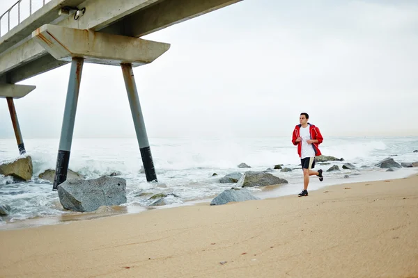 Junger Sportler in weißem T-Shirt und roter Windjacke läuft am Strand entlang und genießt das schöne Meer — Stockfoto