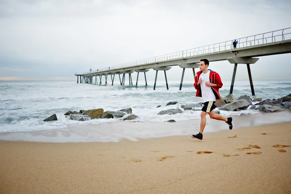 Joven atleta hombre en camiseta blanca corre sobre la arena a lo largo de la playa disfrutando del hermoso mar de la mañana — Foto de Stock