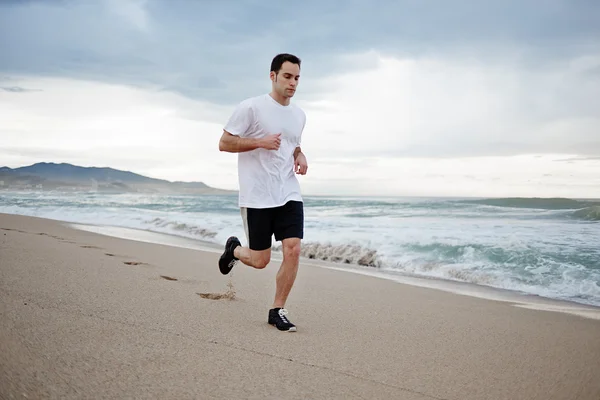 Giovane atleta in t-shirt bianca corre sulla sabbia lungo la spiaggia godendo del bellissimo mare del mattino — Foto Stock