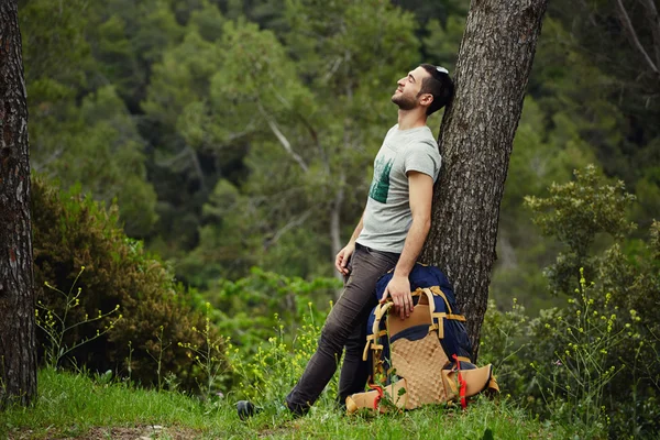 Joven excursionista con mochila apoyada contra un árbol en la pequeña colina de madera — Foto de Stock