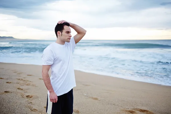 Вмістити чоловіка, який відпочиває після бігу, стоячи на пляжі з морями на фоні — стокове фото