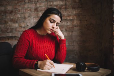 Ciddi bir kadın, kafeteryada kulaklık ve tabletle rahat bir iş yerinde tek başına ders çalışırken not defterine notlar yazıyor.