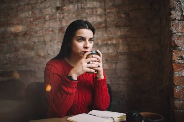 Düşünceli kadın bir bardak sıcak içecekle dinlenirken dirsekleri kafede otururken notlar ve kulaklıklarla gözlerini kaçırıyor.