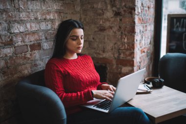 Genç, serbest çalışan, gündelik kıyafetleriyle dizüstü bilgisayarda yazan bir kadın. Küçük bir masada otururken, modern kafede not defteri takıyor.