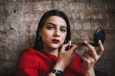 Kusursuz makyajlı, kendine güveni tam genç bir esmer ve kiraz kırmızısı rujlu ve küçük yuvarlak aynalı. Tuğla duvara yaslanırken duygusuz bir şekilde kameraya bakıyor.