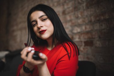 Mükemmel makyajlı, koyu kırmızı rujlu ve elinde küçük yuvarlak aynayla kameraya bakan kendine güvenen genç bir bayan.