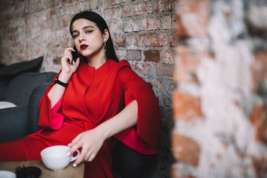 Masanın yanında oturmuş kameraya bakarken akıllı telefondan sohbet eden uzun saçlı, sıcak kahve içen hoş bir kadın.