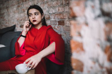 Moda kıyafetleri içinde odaklanmış kadın girişimci elinde kupayla masanın yanında oturuyor ve rahat çalışma ortamında akıllı telefondan sohbet ediyor.