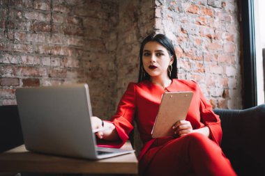 Moda sektöründe yoğunlaşmış bir kadın bilgisayarla sosyal medyada geziniyor ve elinde panoyla masada otururken kameraya bakıyor.