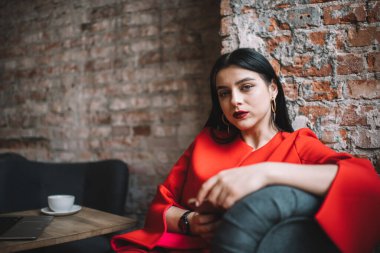 Kırmızı dudaklı düşünceli bir bayan koltuğa yaslanıyor ve geniş bir binada kahve molası sırasında kameraya bakıyor.