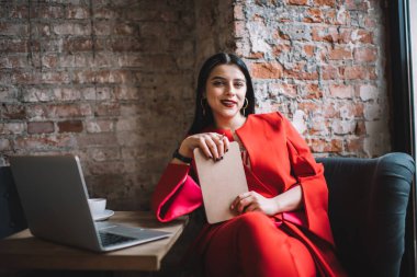 Olumlu kadın tuğla duvarın yanında oturuyor ve çevrimiçi çalışırken önemli bilgileri not alıyor ve kameraya bakıyor.