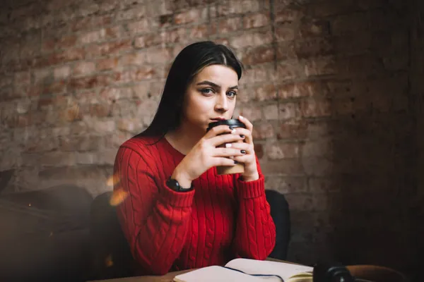 赤いセーターを着た若い女性学生コーヒーを飲みながら 居心地の良いカフェで自由な時間を過ごすノートブックとテーブルに座って — ストック写真