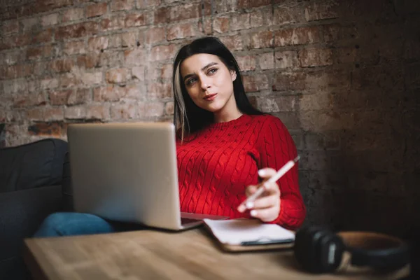Rahat endüstriyel kafe duvarları olan bir masada dizüstü bilgisayar ve not defteriyle otururken gündelik kıyafetlerle yeni fikirler üretmeye çalışan genç yaratıcı kadın.