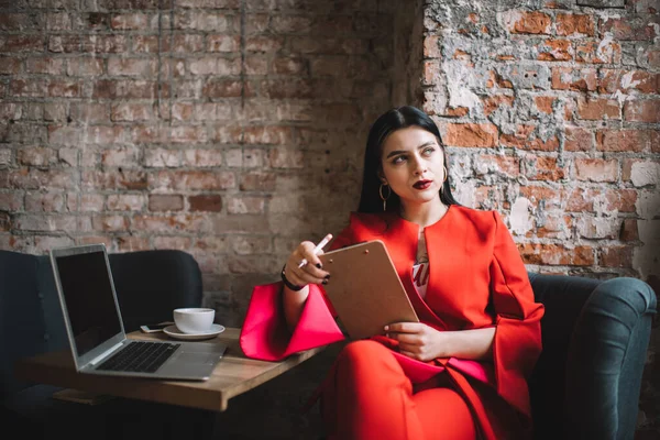 Konsantre olmuş, bacak bacak üstüne atmış bir kadın çalışan elinde panoyla koltukta otururken mali raporlar hakkında kafa yoruyor.