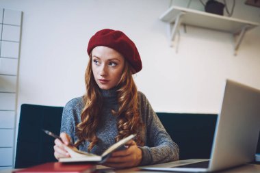 Genç odaklı serbest çalışan kadın iş sırasında not defterinde notlar okurken açık dizüstü bilgisayarla masaya bakıyor.