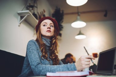 Düşük açılı, düşünceli, serbest çalışan bir kadın yeni bir proje üzerinde çalışırken dizüstü bilgisayarla masaya oturuyor.