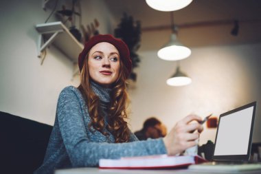 Düşük açılı, kırmızı bereli, rahat giyimli, açık dizüstü bilgisayarı ve not defteriyle masada oturan bir kadın.