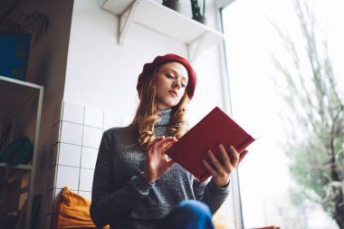 Düşük açılı, uzun saçlı, mavi kazaklı ve kırmızı bereli bir kadın pencerenin yanındaki odada kitap okuyor.