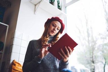 Genç, uzun saçlı, düşünceli, süveter giyen ve bereli roman okuyan bir kadın modern salonda pencerenin yanında oturuyor.