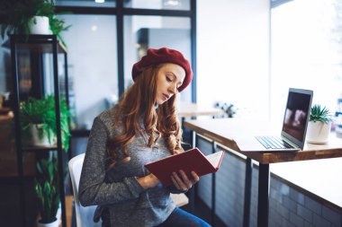 Genç, odaklanmış, mavi kazaklı, kırmızı bereli, masa başında dizüstü bilgisayarı olan bir kadın.