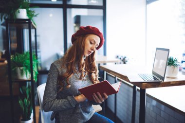 Uzun saçlı, modaya uygun giyinmiş ve kırmızı bereli bir kız öğrenci ders kitabı okuyor. Modern salonda dizüstü bilgisayarla masanın yanında oturuyor.