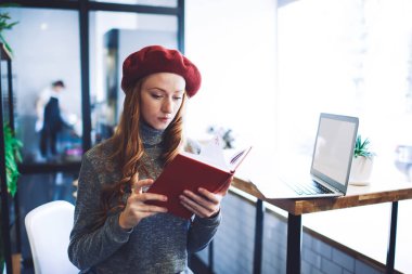 Genç, uzun saçlı, serbest çalışan ve not defterinden bilgi okuyan bir bayan. Masanın yanında dizüstü bilgisayarla modern kafede proje üzerinde çalışıyor.