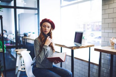 Düşünceli, genç, uzun saçlı, serbest çalışan bir kadın. Masanın yanında oturmuş, dizüstü bilgisayarlı, modern bir kafede oturuyor ve başka tarafa bakıyor.
