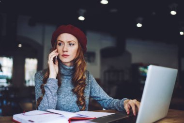 Ciddi kadın uzaktan kumandalı sıradan giysiler içinde masadan uzağa bakıyor dizüstü bilgisayarla oturuyor ve çalışırken telefonla konuşuyor.