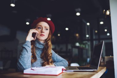 Kafeteryada dizüstü bilgisayar ve not defteriyle otururken uzun saçlı, düşünceli, uzaktan kumandalı, cep telefonlu ve planlama programlı bir kadın.