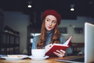 Karizmatik kızıl saçlı, boğazlı kazaklı ve kırmızı bereli kadın kahvaltı sırasında gözlerini başka yöne çevirerek modern kafeye bakarken kırmızı defter kullanıyor.